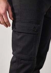 Pantaloni cargo neri con tasca laterale con bottoni, realizzati in tessuto robusto, impreziositi da cuciture visibili e una vestibilità slanciata.