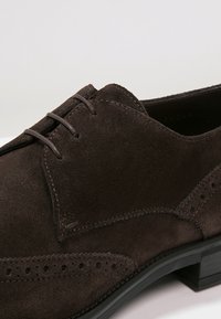 Chaussure à lacets en daim marron avec un détail brogue, un bout arrondi et une semelle en caoutchouc noire. Texture lisse avec des accents de couture contrastants.