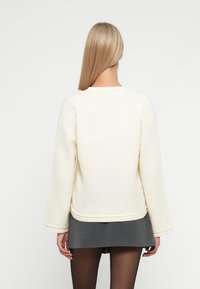 Cremfarbener Pullover mit strukturiertem Strick, Rundhalsausschnitt und Raglanärmeln. Saum und Bündchen mit Rippstrickabschluss. Darunter ein kurzer grauer Rock.