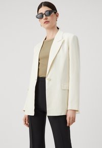 Cream-colored blazer with a single button, notched lapels, and a relaxed fit, paired with a fitted olive top and black pants.