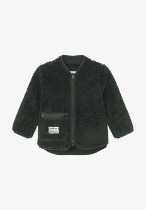 Chaqueta de felpa negra con cremallera, de pie sobre una base de piedra gris, sosteniendo un poste de madera con cables, rodeada de vegetación y una pequeña piscina.
