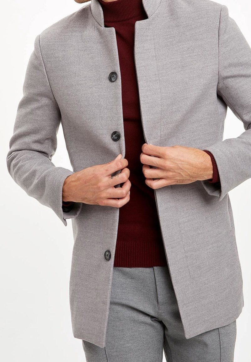 Grå jakke i uldblanding med høj krave, fire sorte knapper og lange ærmer, lagdelt over en bordeaux rullekravesweater.