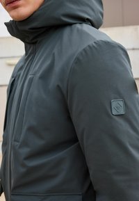 Jachetă izolată de culoare gri închis cu guler înalt, având o peticulă texturată a logo-ului pe brațul stâng și o buzunar frontal. Finisaj neted.