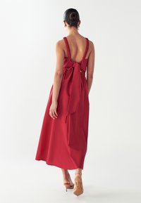 Sieviete, kura stāv ar muguru pret skatītāju, ģērbusies sarkanu midi kleitu bez piedurknēm ar zemu plankumu aizmugurē un lielu sasietu loku, kuru papildina platas stilbenu kurpes ādas krāsā.