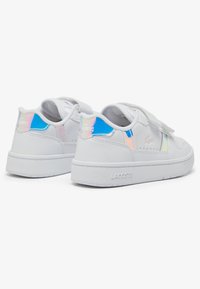Bijele kožne tenisice s glatkom površinom, koje imaju iridescentne naglaske, čičak traku i teksturiranu gumenu potplat. Prisutna je Lacoste logo oznaka.