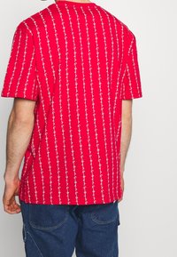 Röd kortärmad skjorta med ett vitt vertikalt mönster av krokar. Tyget verkar slätt med en avslappnad passform och rundad halsringning.