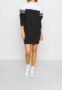 Person som bär en svart och vit långärmad sweatshirtklänning med logotyptext på ärmarna, vita strumpor och vita sneakers mot en enfärgad bakgrund.