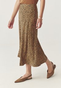 Frau mit einem hellbraunen Midirock mit schwarzen Punkten, braunen spitzen Slingback-Ballerinas und einem goldenen Armband, die vor einem neutralen Hintergrund geht.
