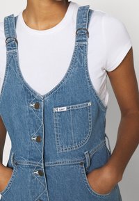 Denimoverall med justerbara axelremmar, knappslå framtill och bröstficka. Ljusblå färg med kontrasterande sömmar och sidofickor.