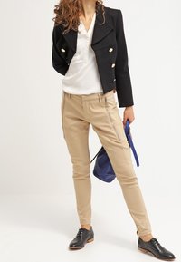 Chaqueta negra cruzada con botones dorados, camisa blanca con cuello, pantalones ajustados color beige con cremalleras y zapatos negros con cordones, sosteniendo una bolsa azul.