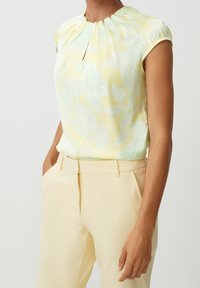 Femme portant un blouse à cap sleeves jaune avec un motif de feuilles bleu clair, rentrée dans un pantalon taille haute jaune pâle, les mains dans les poches.