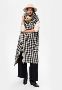 Czarna i biała szal w kratę z frędzlami, narzucony na neutralny top i połączony z czarnymi spodniami oraz botkami na obcasie.