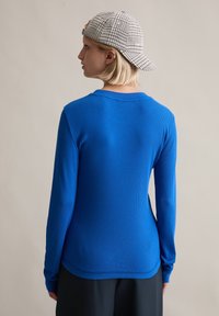 Blaues, geripptes Langarmshirt mit rundem Ausschnitt, kombiniert mit schwarzen Hosen. Eine kariertes Cap wird rückwärts getragen und zeigt ein einfaches Design.
