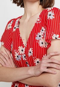 Czerwona sukienka w kwiaty z dekoltem w serek, krótkimi falbaniastymi rękawami i pionowymi paskami. Zauważalne detale to wzory białych i niebieskich kwiatów.