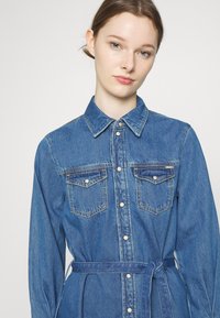 Vestido de camisa de ganga azul com colarinho, fecho frontal com botões, dois bolsos no peito e um cinto correspondente na cintura. Textura suave e corte descontraído.