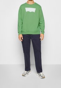 Une personne porte un sweat-shirt vert Levi's, un pantalon bleu foncé et des baskets blanches et grises, se tenant contre un fond gris clair uni.