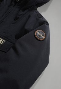 Mörk marinblå jacka med huva, slät textur och en cirkelformad logotyp i orange och svart på ärmen. Inkluderar en frontdragkedja.