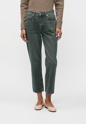 Personne portant un jean droit gris foncé taille haute, un pull beige et des ballerines rose clair, debout devant un fond uni.