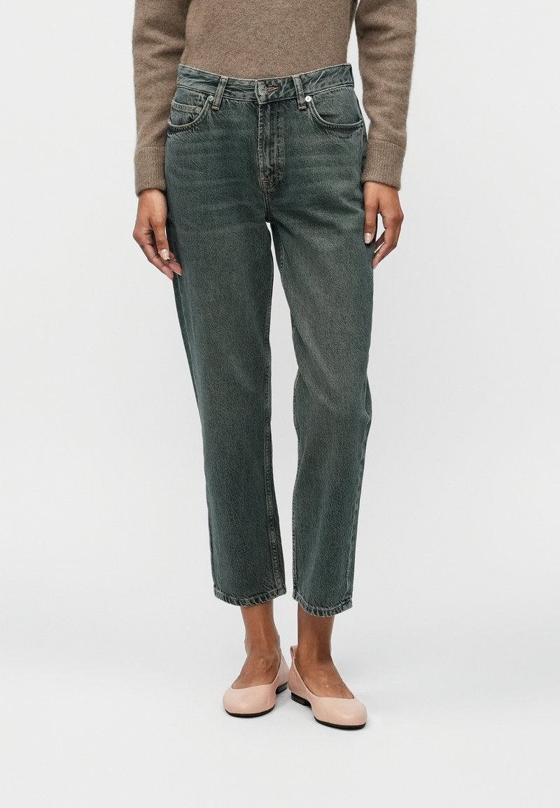 Personne portant un jean droit gris foncé taille haute, un pull beige et des ballerines rose clair, debout devant un fond uni.