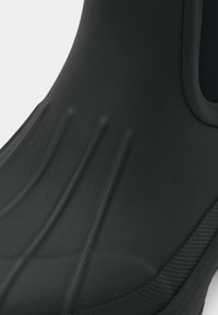 Botte en caoutchouc noir avec une surface texturée, présentant des lignes verticales pour l'adhérence et une doublure intérieure lisse. Le design comprend un haut arrondi.