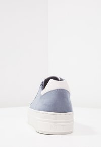 Baskets en daim gris avec languette de talon blanche et semelle épaisse en caoutchouc blanc, vues de dos sur une surface blanche avec un fond blanc uni.