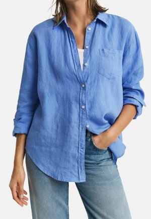 Kvinde iført en løstsiddende blå skjorte med knapper, med ærmerne rullet op og lyseblå jeans, den ene hånd i lommen, hvid undertrøje synlig.