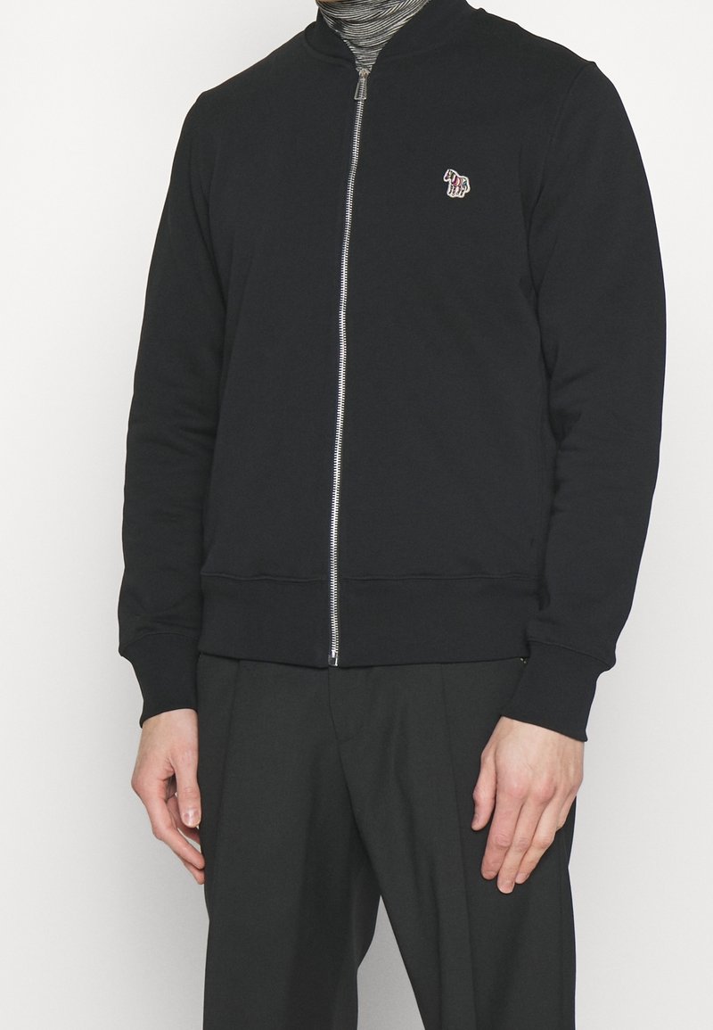 Sweat-shirt noir à fermeture éclair avec col ajusté, poignets côtelés et texture semblable à la laine. Présente un petit motif brodé sur la poitrine.