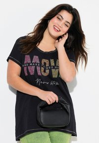 Camiseta negra de manga corta, con letras texturizadas en multicolor. Combinada con pantalones verdes y un bolso negro texturizado.