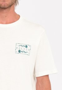 White short-sleeve t-shirt with a teal graphic print on the left chest featuring the word "VOLCOM" and floral accents. Soft cotton material.