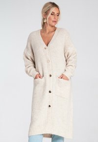 Kobieta z blond włosami, ubrana w długi, beżowy kardigan z dużymi guzikami i kieszeniami, w połączeniu z jasnoniebieskimi dżinsami, stojąca na białym tle.