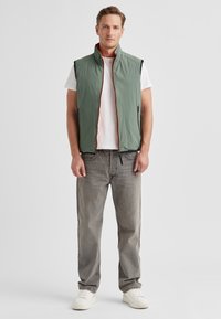 Green lightweight vest with high collar, zip front, and orange accents. Worn over a white t-shirt with gray, relaxed-fit jeans and white sneakers.