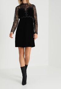 Robe en velours noir avec des manches longues en dentelle transparente et un décolleté festonné, présentant un corsage ajusté et une jupe évasée.