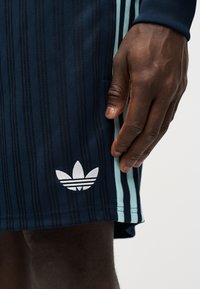 Sötét bőrű kéz pihen egy tengerészkék rövidnadrág mellett, amely világoskék csíkokkal és a combon fehér Adidas Trefoil logóval van díszítve.