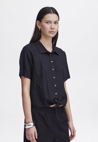 Camicetta nera a maniche corte realizzata in un tessuto leggero, con colletto, chiusura frontale con bottoni e dettaglio annodato sul davanti. Indossata con pantaloni coordinati.