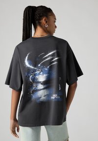 T-shirt preta de algodão com uma grande imagem gráfica nas costas, representando um céu noturno com estrelas e uma frase em texto azul, mangas curtas.