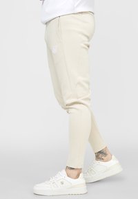 Béžové joggers vyrobené z měkké látky s tapered designem, mají boční kapsy a jemné výšivky loga na stehně. V kombinaci s bílými teniskami.