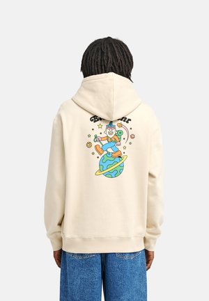 Personne portant un sweat à capuche crème avec un sorcier de dessin animé coloré marchant sur une planète avec un motif d’étoiles et de planètes dans le dos.