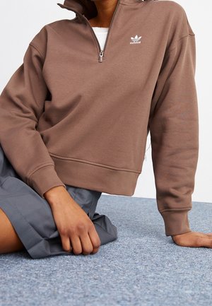 Person sitzt auf blauem Teppich, trägt braunen Adidas-Quarter-Zip-Pullover und grauen Rock, rechte Hand auf dem Boden, linke Hand auf dem Knie.