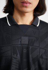 Polo preto da Nike com padrão texturizado, com acabamento branco na gola e um patch do logo Nike Air no peito, apresentando um design geométrico.