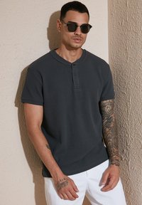 Maglietta henley grigia scura a maniche corte con tessuto strutturato a coste, dotata di una patta con bottoni e maniche arrotolate. Indossata con pantaloni bianchi.