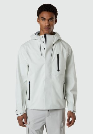 Mann trägt eine hellgraue wasserdichte Jacke mit Kapuze und schwarzen Reißverschlussdetails, steht vor einem einfarbigen hellgrauen Hintergrund.