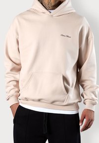 Beżowa bluza z kapturem z przednią kieszenią, ściągaczami na rękawach i dole; na piersi małe czarne logo. Miękka tekstura tkaniny.