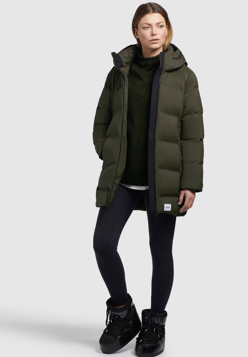 Dunkelgrüner gepolsterter Mantel mit Kapuze, Frontreißverschluss und schwarzem Besatz. Trägt einen dunklen Pullover und schwarze Leggings mit schwarzen Stiefeln.