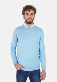 Maglione azzurro chiaro lavorato a maglia con scollo rotondo, maniche lunghe e polsini a costine. Indossato sopra una camicia bianca con colletto, abbinato a jeans scuri.