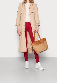 Mujer con abrigo trench beige, suéter blanco, pantalones rojos, zapatillas blancas, sosteniendo un bolso de mimbre color marrón claro con detalles de cuero.