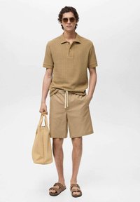 Polo shirt a coste marrone chiaro, pantaloni corti abbinati, sandali marroni e borsa a tracolla intrecciata. Presenta una vestibilità comoda e uno stile casual.