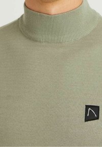 Pull col roulé côtelé vert olive avec une finition texturée et un petit patch noir arborant un accent graphique blanc.