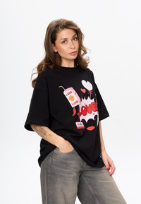 Czarny oversize T-shirt z bawełny z kolorową grafiką przedstawiającą napój, serca i słowo "LOVE". W zestawieniu z jasnoszarymi spodniami.