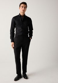 Camicia nera con bottoni e maniche lunghe, abbinata a pantaloni neri su misura e scarpe nere con trama. Design semplice, elegante e monocromatico.