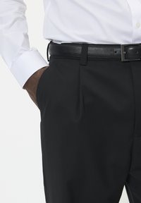 Czarne, dopasowane spodnie o gładkiej fakturze, z pojedynczą przednią kieszenią i czarnym skórzanym paskiem. Widoczny mankiet białej koszuli.
